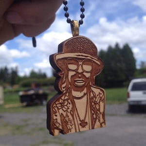 May include: A wooden pendant necklace featuring a laser-cut image of a person wearing a hat, sunglasses, and a floral shirt. The pendant is brown and has a gold chain.