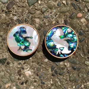 May include: Two round wooden coasters with a white silhouette of a person sitting on a chair. The coasters are made of abalone shell with iridescent green, blue, and purple colors.