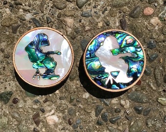 Puede incluir: Dos posavasos redondos de madera con una silueta blanca de una persona sentada en una silla. Los posavasos están hechos de concha de abulón con colores iridiscentes verdes, azules y morados.