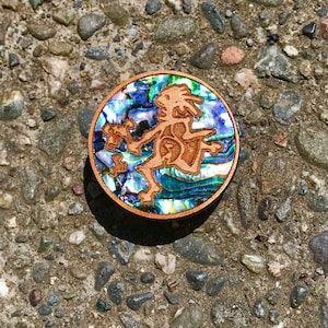 May include: A round wooden pin with a blue, green, and iridescent abalone shell inlay. The pin features a carved wooden figure of a dancing person.