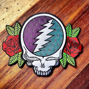 May include: Embroidered patch featuring a skull with a purple and teal circle design and a white lightning bolt. Red roses and green leaves flank the skull. The patch is on a wooden surface.