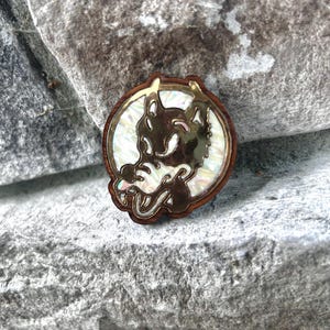 May include: A round, wooden pin with a gold-colored outline and a detailed wolf design. The wolf is set against a pearlescent background. The pin is a unique accessory.