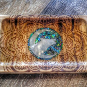 May include: Rectangular wooden tray with a carved mandala design. The center features a wolf silhouette within a crescent moon, inlaid with iridescent abalone shell. The tray is made of light-colored wood.