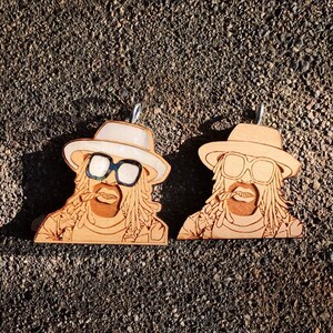 May include: Two wooden pendants featuring a stylized portrait of a man with dreadlocks, sunglasses, and a hat. The pendants are light brown with engraved details, and each has a small metal loop at the top for hanging. The background is a textured, dark surface.