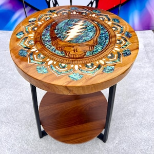 May include: A round wooden side table with a glossy finish, featuring a central skull graphic with a lightning bolt inlay surrounded by iridescent blue and green abalone shell accents in a mandala pattern, supported by black metal legs.