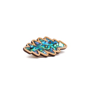 May include: A wooden brooch in the shape of a stylized leaf. The brooch features a vibrant abalone shell inlay with iridescent blues, greens, and purples. The wooden frame has a light brown color.
