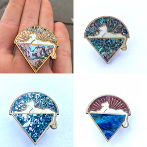 May include: Four different variations of a wooden pin featuring a white silhouette of a cat lying down on a bed of iridescent abalone shell. The cat is silhouetted against a sunburst design. The pins are in a triangular shape.