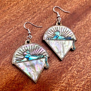 May include: Wooden earrings with a fan-shaped design, featuring a mother-of-pearl inlay and a stylized cat motif. The cat is depicted in shades of blue and teal, resting on a platform. The earrings have silver-colored ear wires.