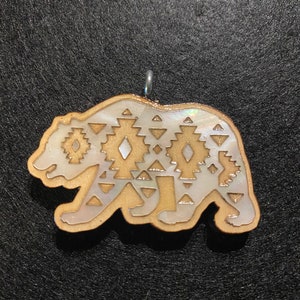 Puede incluir: Un colgante de oso de madera con un patr&oacute;n geom&eacute;trico en blanco y dorado. El oso est&aacute; tallado en madera y tiene un lazo plateado para colgar.