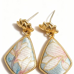 May include: A pair of gold-coloured earrings. Each earring features a gold lotus flower stud and a teardrop-shaped pendant. The pendant has a light blue background with a floral pattern in white and pink, framed by a gold border.