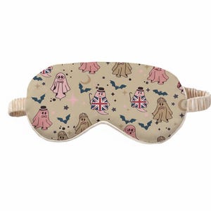May include: A beige sleep mask featuring a repeating pattern of cartoon ghosts, bats, stars, and moons. Some ghosts are adorned with hats and Union Jack flags. The mask has a beige elasticated band.