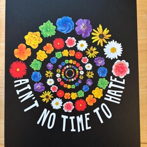 May include: A black print featuring a spiral of colorful flowers in shades of red, orange, yellow, green, blue, and purple. The words "Ain't No Time To Hate" are in white, arched around the flowers.