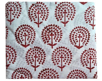 May include: White fabric with a repeating pattern of red floral designs. Each design features a central cross-like shape surrounded by a circular, gear-like pattern. The fabric is folded, showing the pattern's repetition.
