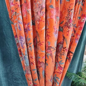 May include: Orange floral patterned curtains. The curtains feature a detailed design of flowers and birds in shades of pink, purple, and blue. The fabric has a velvet-like texture, adding depth to the design. The curtains are hanging in vertical folds.