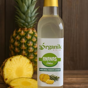 以下が含まれることがあります： 金色のキャップが付いた透明なガラス瓶で、「Organik Evim Ananas Sirkesi」とラベルが貼られています。丸ごとのパイナップルとスライスされたパイナップルの隣にあります。緑と黄色のテキストのラベルには、パイナップルのイラストが含まれています。背景は木製の表面です。