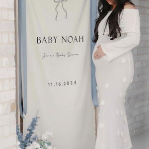 May include: A white banner with the text "Celebrating BABY NOAH Jane's Baby Shower 11.16.2024" is displayed. A woman in a white off-the-shoulder dress with floral accents stands next to the banner. Blue and white flowers are at the base of the banner.