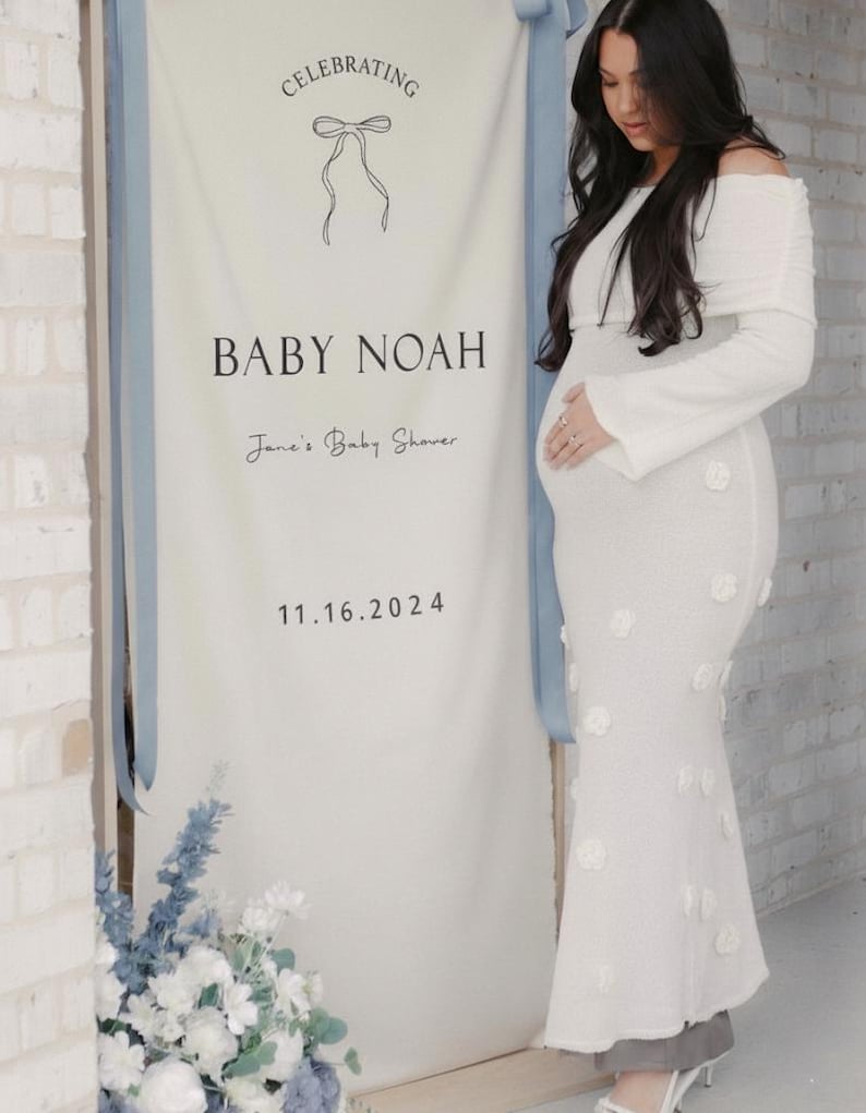 May include: A white banner with the text "Celebrating BABY NOAH Jane's Baby Shower 11.16.2024" is displayed. A woman in a white off-the-shoulder dress with floral accents stands next to the banner. Blue and white flowers are at the base of the banner.