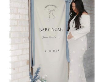 May include: A white banner with blue ribbons and the text "Celebrating Baby Noah" for a baby shower. The date 11.16.2024 is also displayed. A woman in a white dress stands next to the banner.