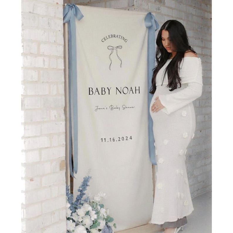 May include: A white banner with blue ribbons and the text "Celebrating Baby Noah" for a baby shower. The date 11.16.2024 is also displayed. A woman in a white dress stands next to the banner.