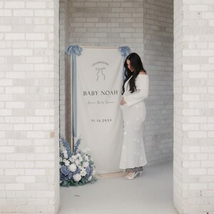 May include: A white banner with the text "CELEBRATING BABY NOAH" and the date 11.16.2024, decorated with blue ribbons and flowers. A woman in a white dress stands near the banner.