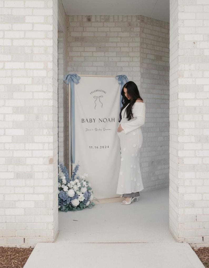 May include: A white banner with the text "CELEBRATING BABY NOAH" and the date 11.16.2024, decorated with blue ribbons and flowers. A woman in a white dress stands near the banner.