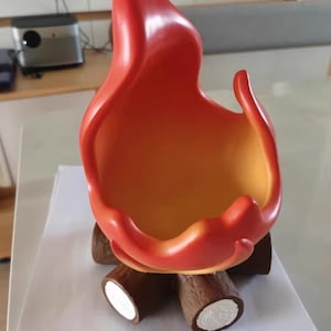 以下が含まれることがあります： 赤とオレンジの色合いの、炎の形をした装飾的なボウル。茶色の丸太型の支えの上に置かれています。ボウルの内側は温かみのある黄オレンジ色で、鮮やかなコントラストを生み出しています。デザインはキャンプファイヤーに似ています。