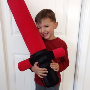 Puede incluir: Un niño sostiene un gran juguete de sable de luz de peluche rojo y negro. El sable de luz es aproximadamente de la misma altura que el niño. El niño sonríe y mira a la cámara.