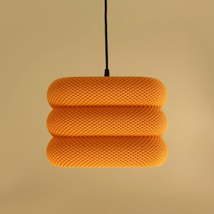Könnte beinhalten: Orangefarbene Pendelleuchte mit strukturierter, wabenartiger Oberfläche. Die Leuchte hat ein gestapeltes, zylindrisches Design und hängt an einem schwarzen Kabel. Die Leuchte hat eine warme Farbe.
