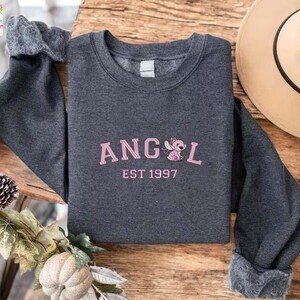 A gray sweatshirt with the text "ANGEL EST 1997" printed on it, along with a pink cartoon character, is displayed on a wooden surface.