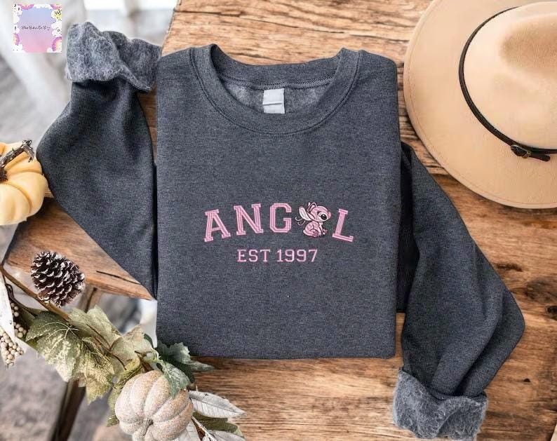 A gray sweatshirt with the text "ANGEL EST 1997" printed on it, along with a pink cartoon character, is displayed on a wooden surface.