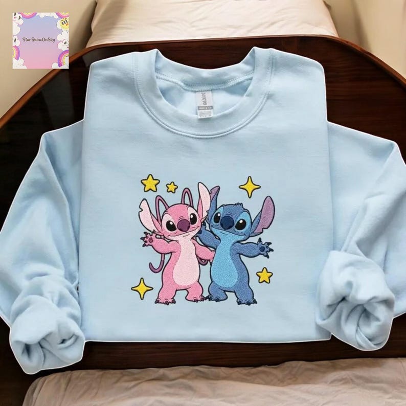 A light blue sweatshirt with a cartoon character design featuring two cats, one pink and one blue, surrounded by stars.