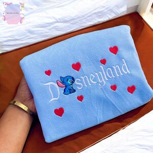 A hand holding a blue towel with the word "Disneyland" and a small blue character embroidered on it, surrounded by red heart shapes.