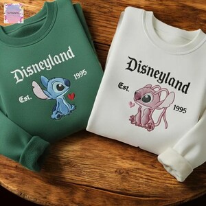 two sweatshirts with Disneyland branding, one green and one white, both with the text "Disneyland Est. 1995" and the Disney character Lilo and Stitch.
