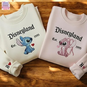two Disney-themed sweatshirts with the character Lilo and the character Stitch, both from the Disney movie Lilo & Stitch. The sweatshirts are displayed on a wooden surface, with the left sweatshirt being white and the right sweatshirt being pink.