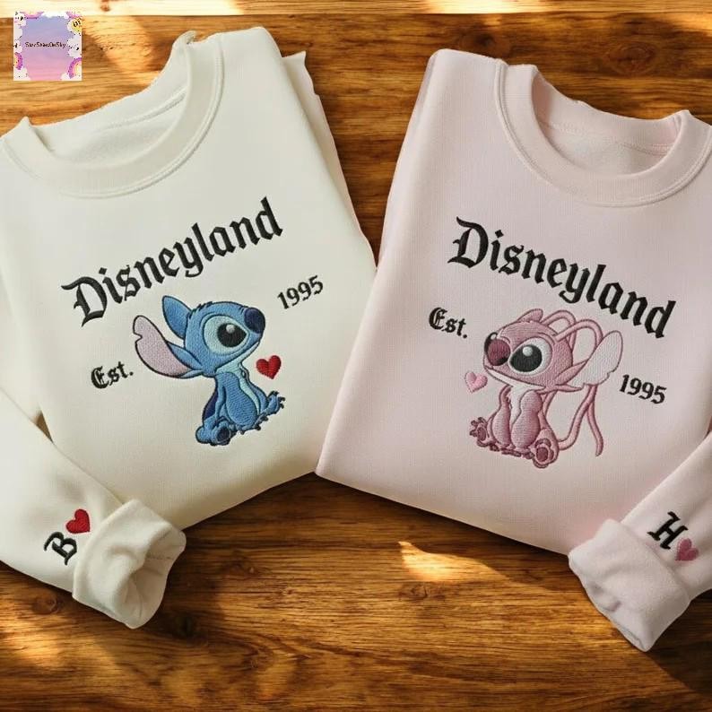 two Disney-themed sweatshirts with the character Lilo and the character Stitch, both from the Disney movie Lilo & Stitch. The sweatshirts are displayed on a wooden surface, with the left sweatshirt being white and the right sweatshirt being pink.
