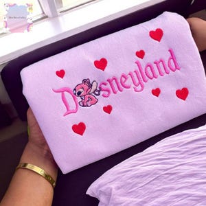 A person is holding a pink towel with the word "Disneyland" embroidered on it, surrounded by red hearts.
