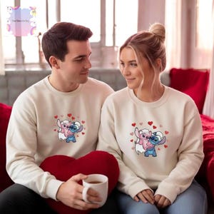 A man and a woman are sitting together on a couch, wearing matching white sweatshirts with a cartoon character design.