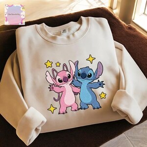 A white sweatshirt with a colorful embroidered design featuring two cartoon characters, a pink alien and a blue alien, surrounded by stars and a heart.