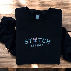 A black sweatshirt with the word "STITCH" and "EST 1999" printed on it, along with a small Stitch character embroidered on the front.