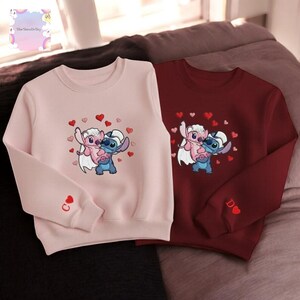 two sweatshirts with cartoon characters from the Disney movie Lilo & Stitch. The sweatshirt on the left is pink, and the one on the right is burgundy. Both sweatshirts have a heart design and the character Lilo & Stitch hugging each other.