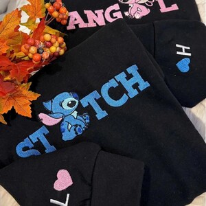a black sweatshirt with the word "STITCH" embroidered on it, along with a character from the Disney movie Lilo and Stitch. The sweatshirt is placed on a bed with a floral arrangement and autumn leaves.