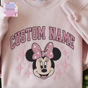 A pink sweatshirt with a Minnie Mouse design and the text "CUSTOM NAME" printed on it.