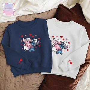 two sweatshirts with cartoon characters from the Disney movie Lilo & Stitch. The sweatshirt on the left is navy blue and the one on the right is white. Both sweatshirts have a heart design and the characters are depicted in a playful, affectionate manner.
