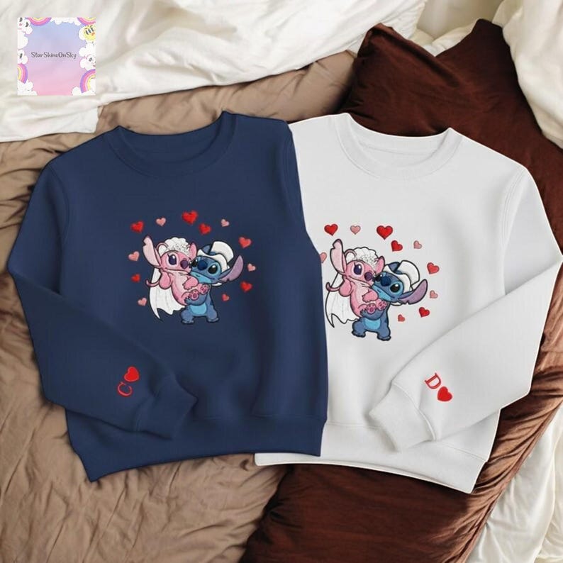 two sweatshirts with cartoon characters from the Disney movie Lilo & Stitch. The sweatshirt on the left is navy blue and the one on the right is white. Both sweatshirts have a heart design and the characters are depicted in a playful, affectionate manner.