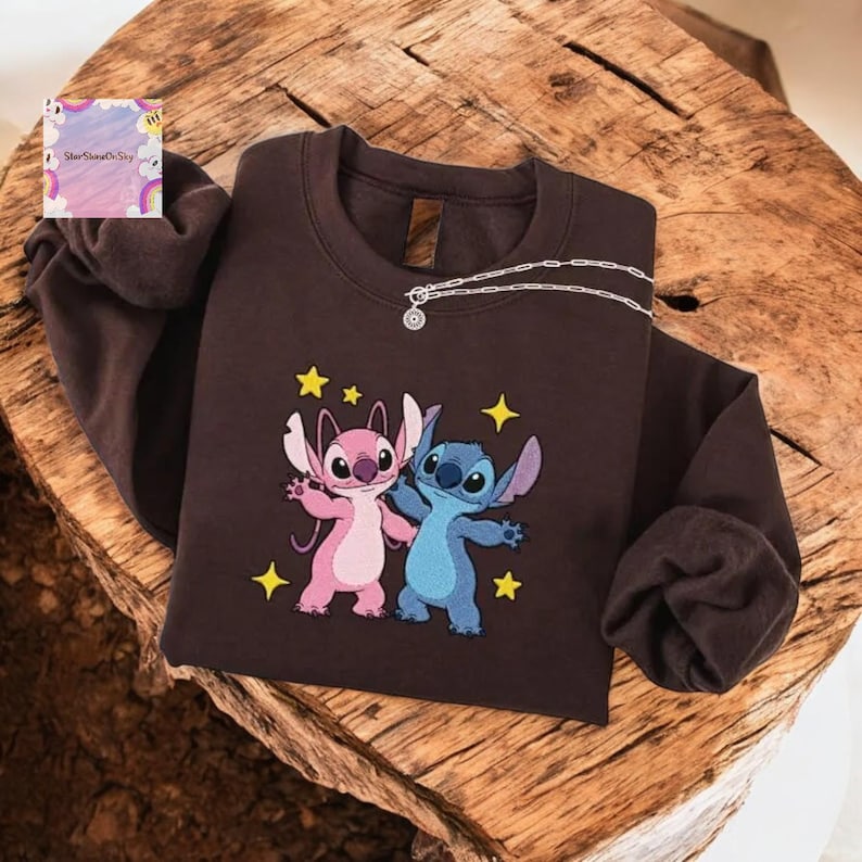 A brown sweatshirt with a cartoon character design, featuring two characters - one pink and one blue - standing together and surrounded by stars.