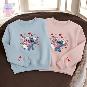 Two children's sweatshirts with a character design, featuring a blue character and a pink character surrounded by hearts.