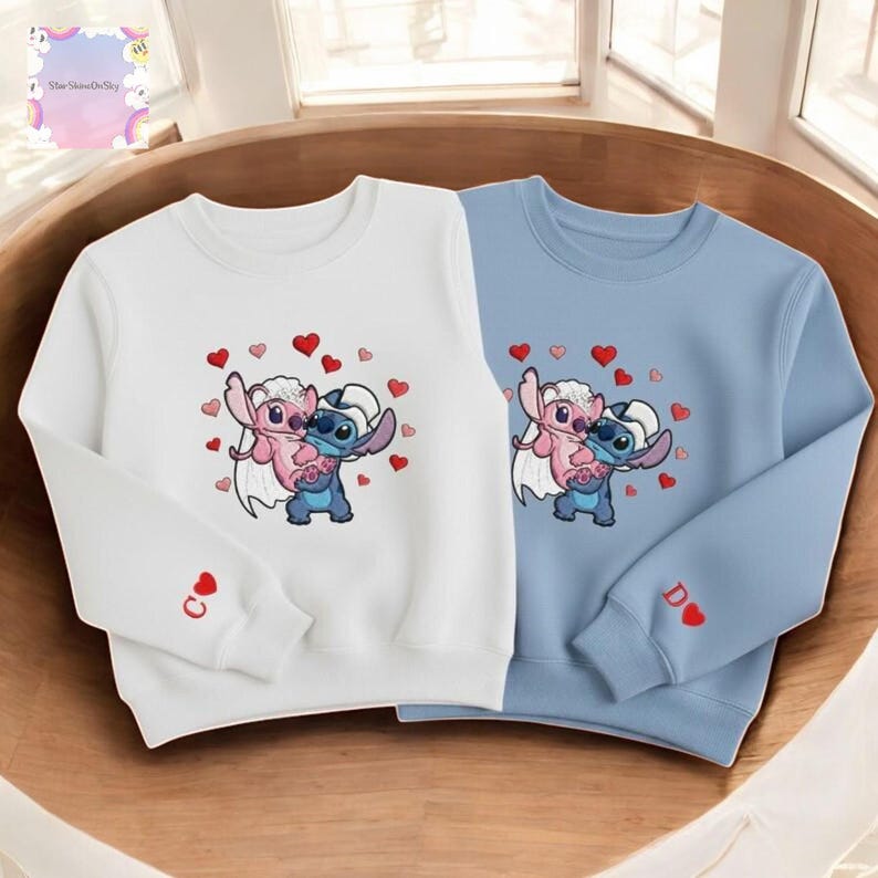 two sweatshirts with a cartoon character design, each with a heart motif. The sweatshirt on the left is white, while the one on the right is blue.