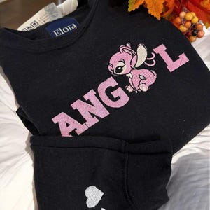 A black sweatshirt with a pink embroidered character and the word "ANGEL" printed on it, along with a small white heart symbol. The sweatshirt is placed on a bed, and there are some orange leaves visible in the background.