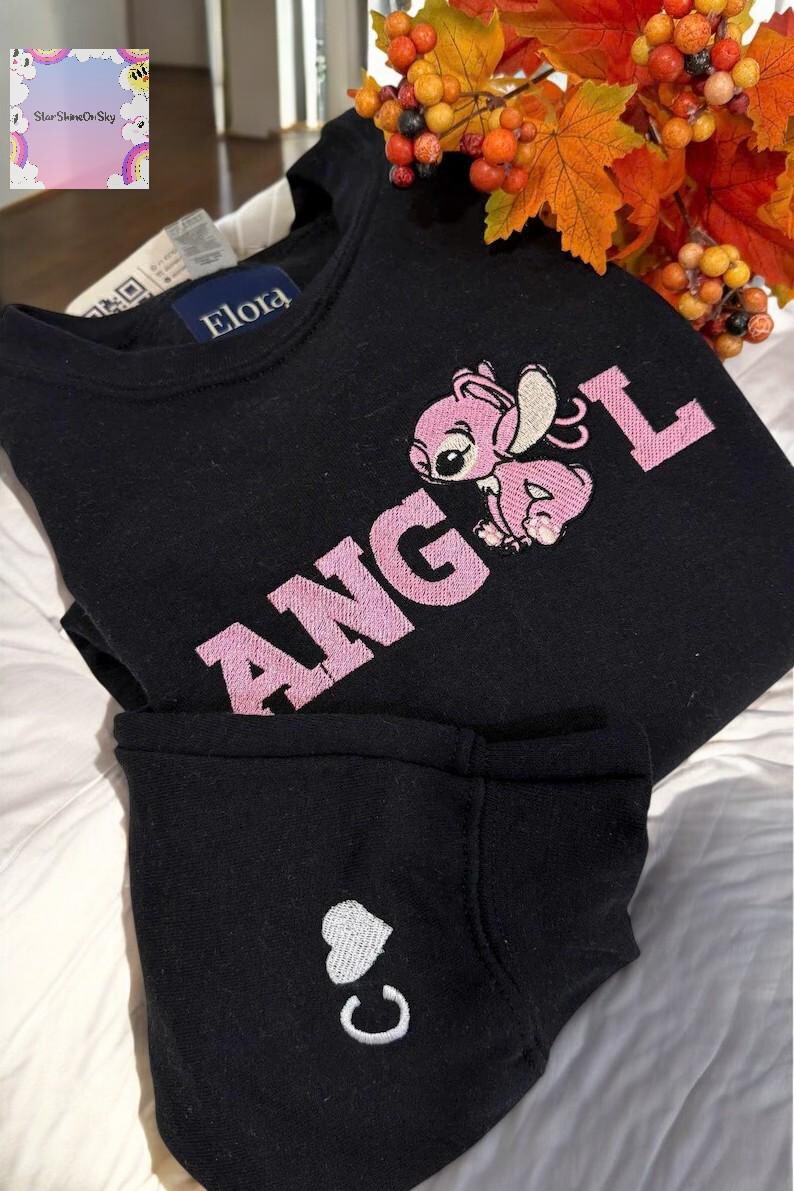 A black sweatshirt with a pink embroidered character and the word "ANGEL" printed on it, along with a small white heart symbol. The sweatshirt is placed on a bed, and there are some orange leaves visible in the background.