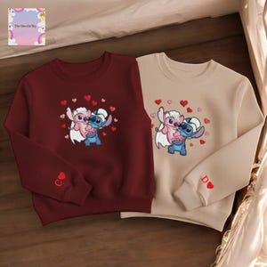 two sweatshirts with the same cartoon character design, featuring a pink and blue character surrounded by hearts.